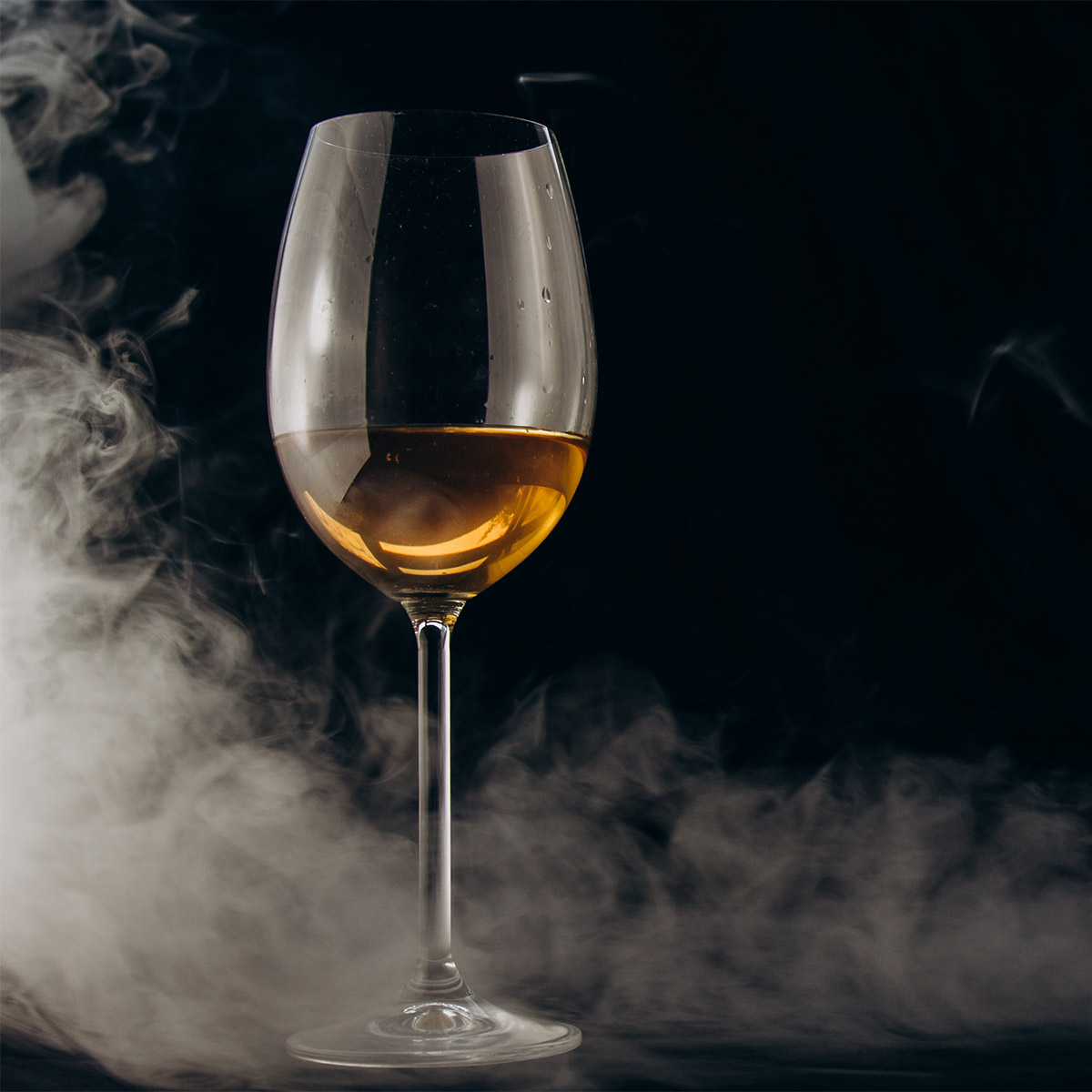 A glass of white wine on a black background as smoke envelops the glass