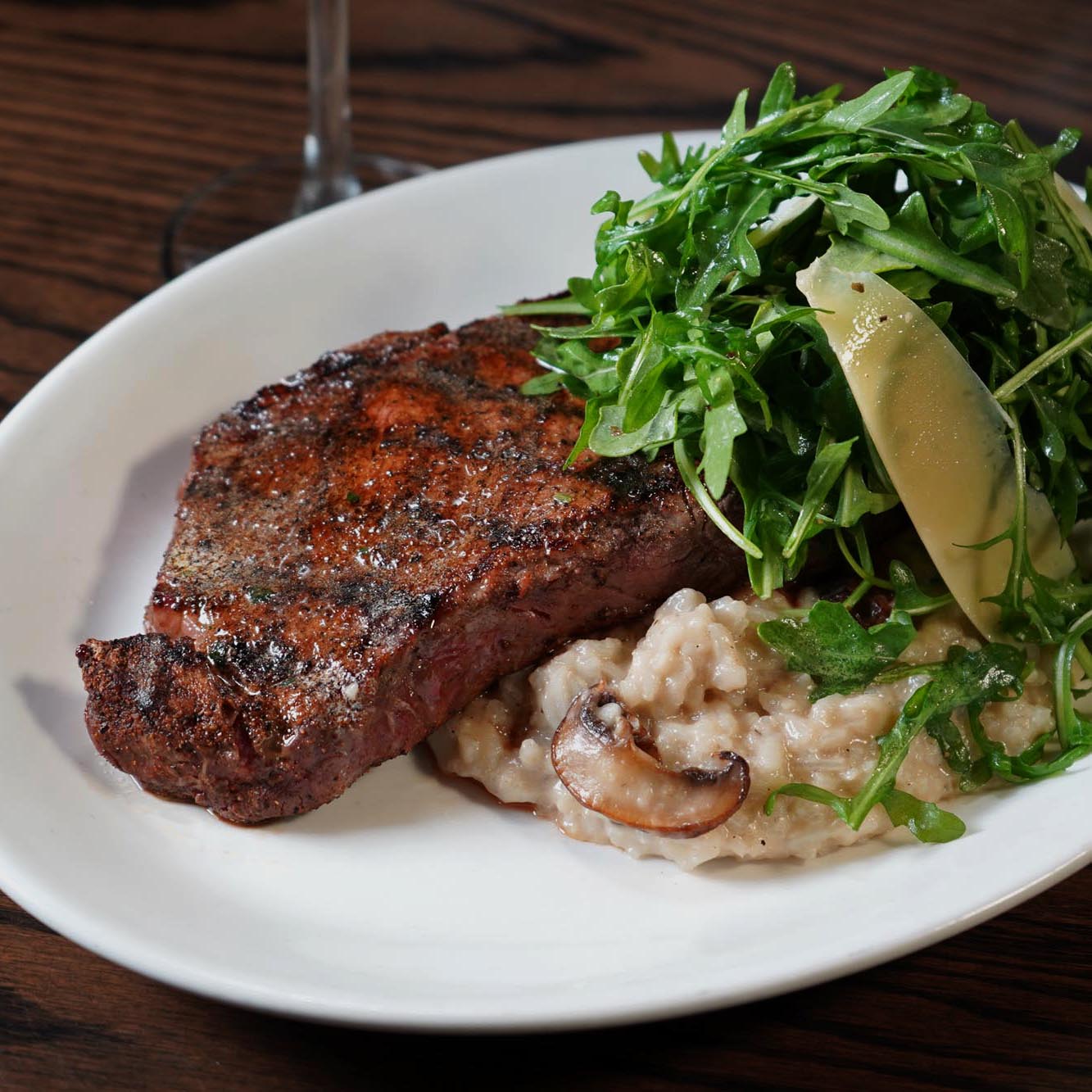 A Wagyu New York Strip steak plated with mushroom risotto and arugula salad.