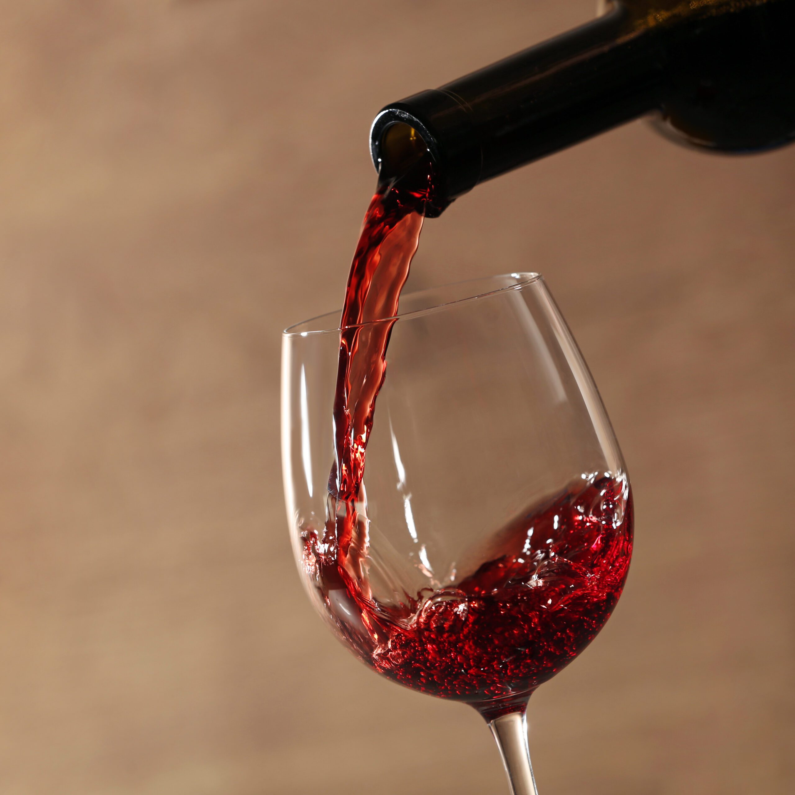 Pouring red wine into glass from bottle against blurred beige background, closeup.